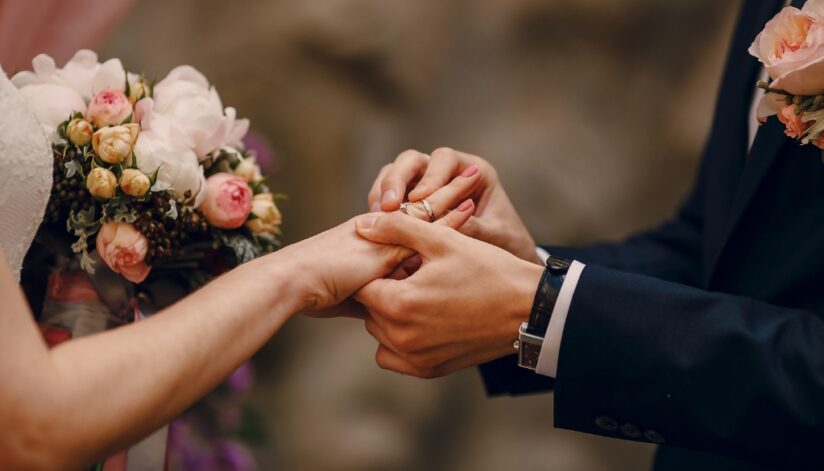 A bride places a wedding ring on the groom’s hand during the ceremony while holding a bouquet of pink and white flowers.