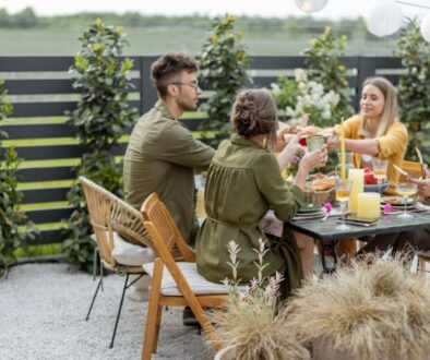 Four friends sit at an outdoor table sharing food and drinks among greenery and string lights.