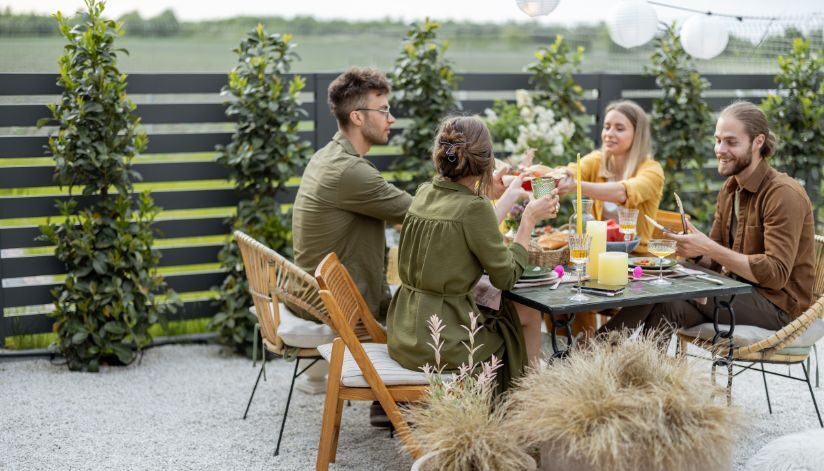 Four friends sit at an outdoor table sharing food and drinks among greenery and string lights.