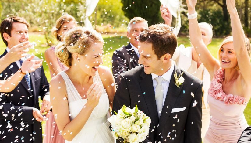 A smiling bride and groom are showered with confetti by their guests outdoors.