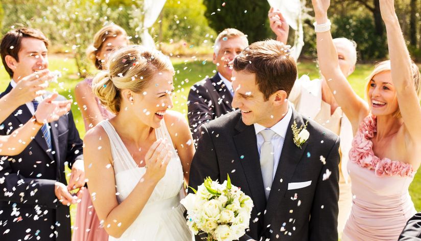 A smiling bride and groom are showered with confetti by their guests outdoors.