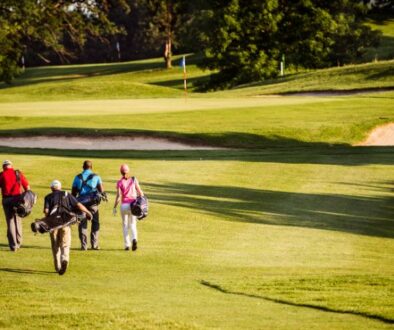 Four golfers with bags walk across the sunny green fairway.