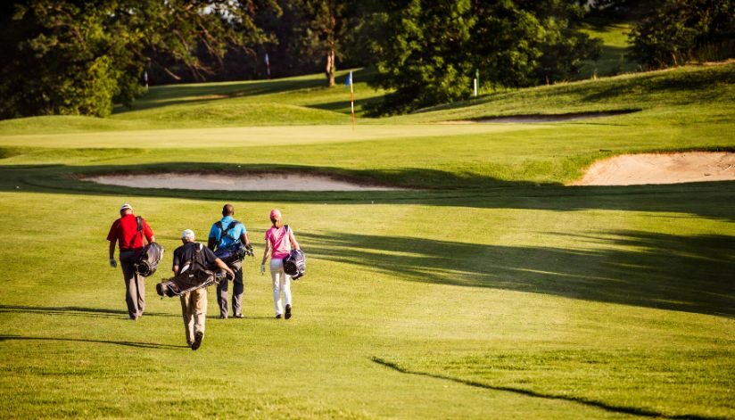 Four golfers with bags walk across the sunny green fairway.