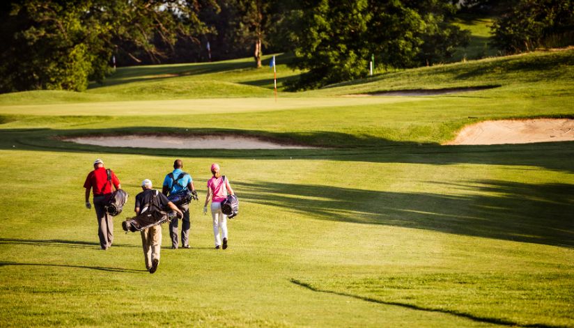 Four golfers with bags walk across the sunny green fairway.