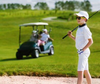 A smiling boy in a white golf outfit stands with his club on the green.