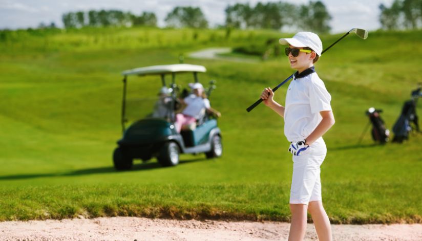 A smiling boy in a white golf outfit stands with his club on the green.