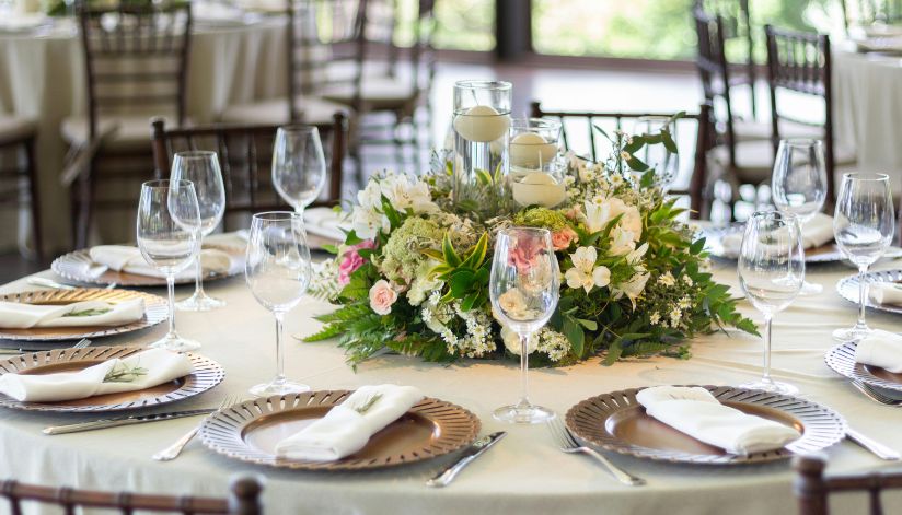 A round table formally set with glassware, folded napkins, and a floral centerpiece.