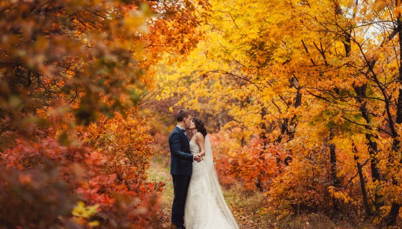 A bride and groom embrace and kiss on a path at Drugan’s Castle Mound, framed by vibrant red, orange, and yellow autumn trees.