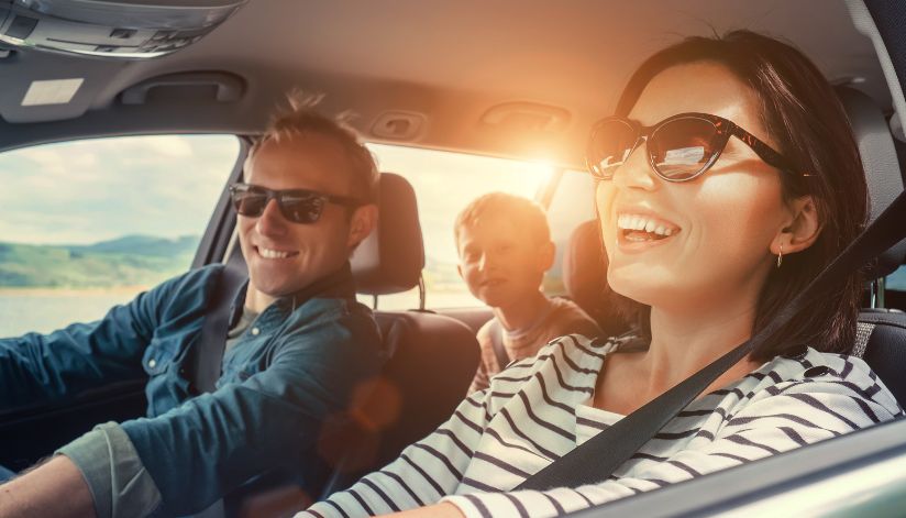 A smiling family wearing sunglasses drives past Drugan’s Castle Mound in Holmen, Wisconsin, sunlight streaming in the car.
