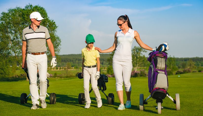 A family of three, each carrying a golf bag and dressed in sporty attire, walks across the green at Drugan’s Castle Mound in Holmen, WI.